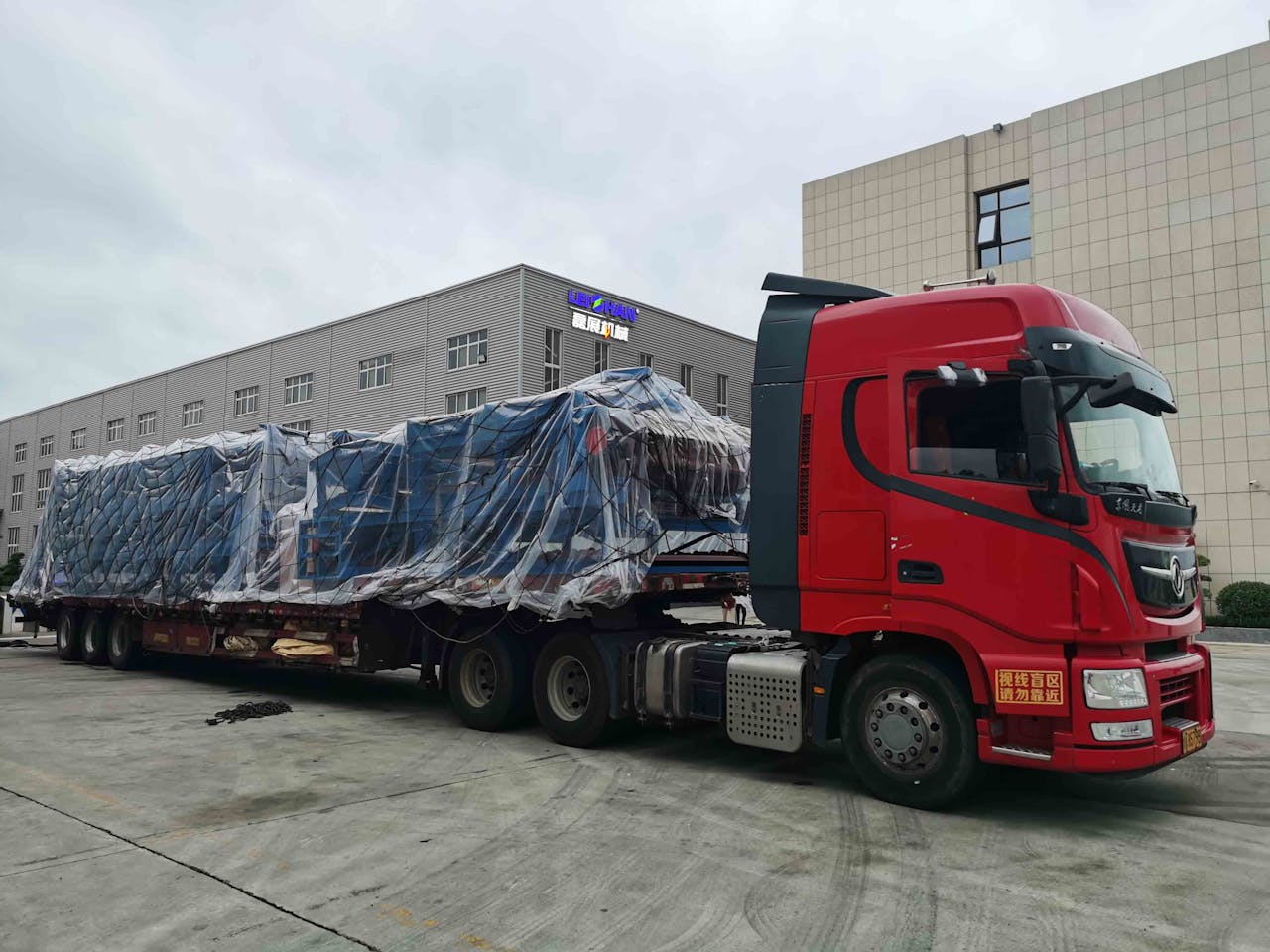 Home Red truck with large covered cargo in an industrial area, overcast day.