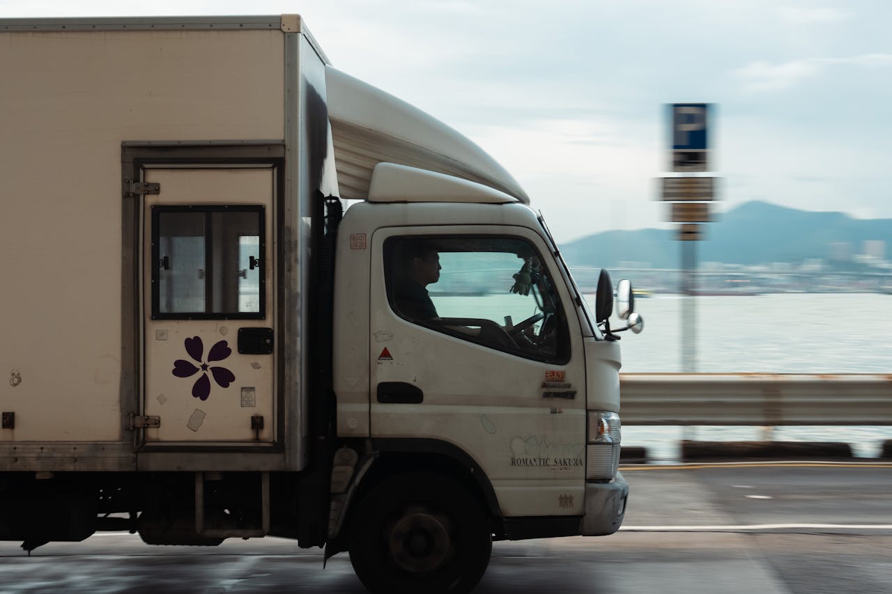 Services A delivery truck driving along a coastal road with a mountain backdrop on a clear day.